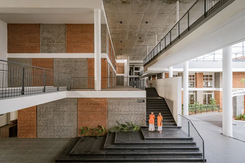 Interior staircase with black treads ascending past perforated brick panels and two figures in orange robes