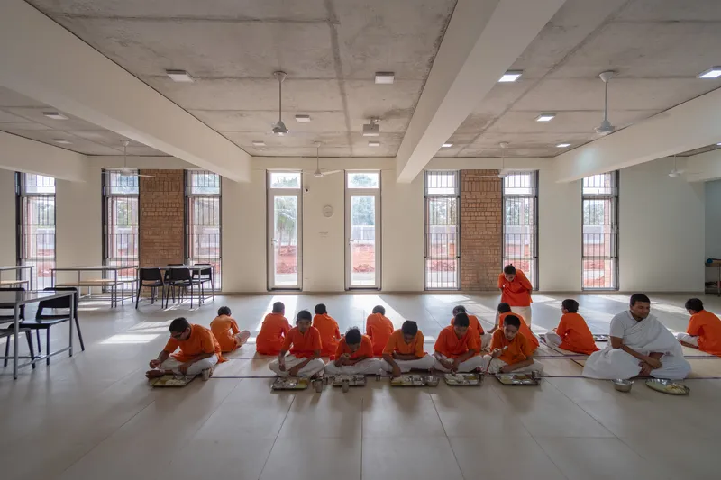 Interior dining hall with exposed concrete ceiling where students in orange robes sit for a meal
