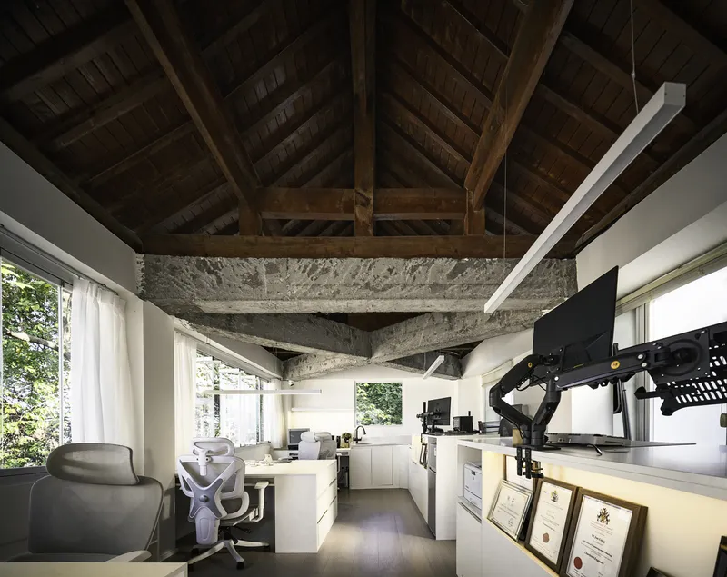 Office interior with exposed timber roof trusses and rough concrete beam above white desks