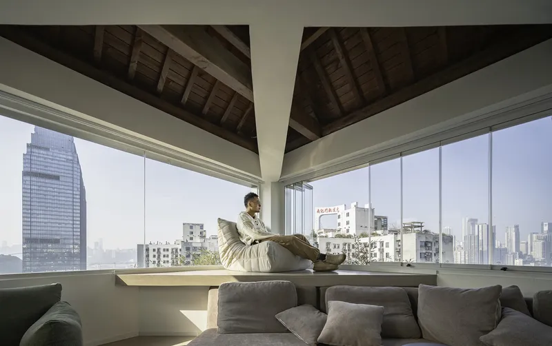 Window seat beneath crossed timber roof planes with cityscape view and seated figure reading