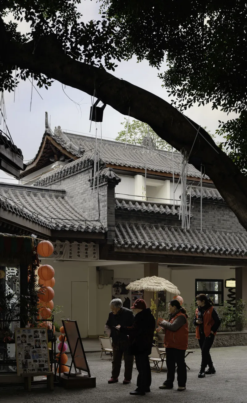 View from beneath a large tree framing a traditional tiled-roof pavilion with visitors gathered in the courtyard
