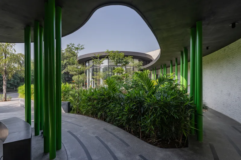 Covered walkway with green tubular columns framing planted beds and the cylindrical glass pavilion with curving canopy