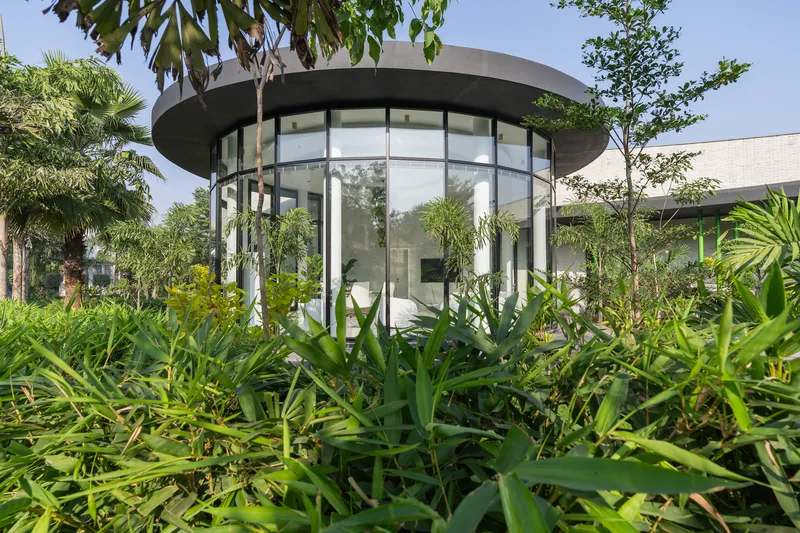 Cylindrical glass pavilion with concrete canopy surrounded by dense tropical plantings under midday sun