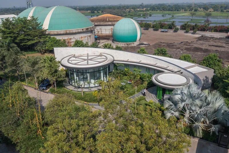 Aerial view of the curved structure with circular volumes among palms and green domed buildings beyond a waterway