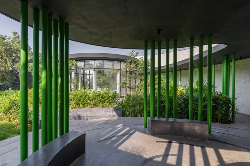 Covered terrace with green tubular columns casting afternoon shadows across concrete benches and planted beds