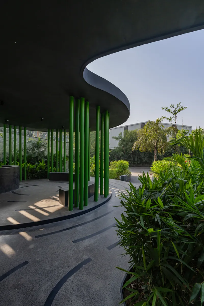 Circular pavilion with bright green cylindrical columns beneath a dark soffit casting shadows on stone paving