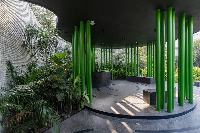 Open-air pavilion interior with green tubular columns, planted beds, and a grey brick wall