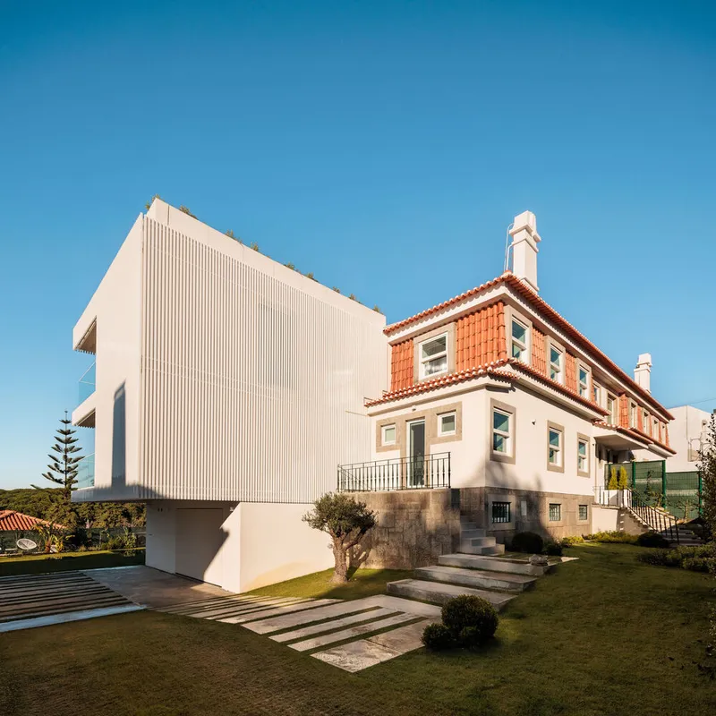 Side elevation showing the ribbed white volume adjacent to an older terracotta-roofed house and lawn