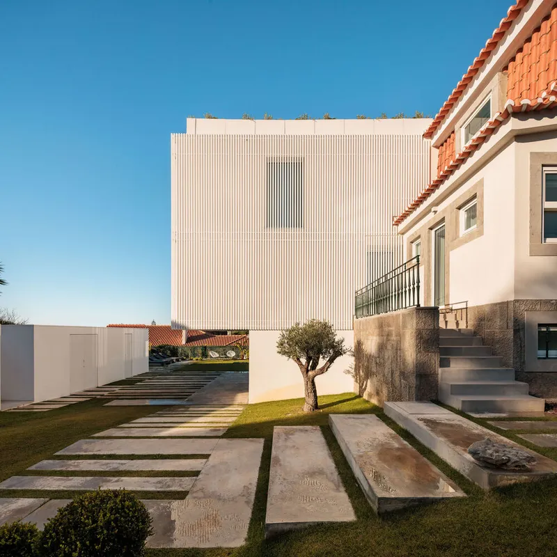 Garden view showing vertical louvered facade beside an orange tile-roofed house with stone pavers crossing lawn