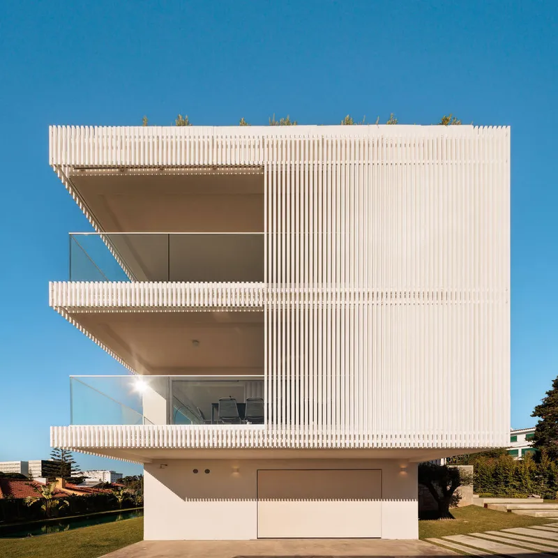 Street facade showing three stacked balconies with glass railings framed by vertical white louvers under blue sky