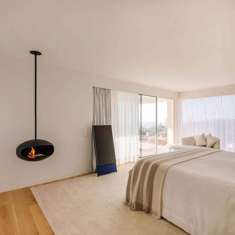 Bedroom with suspended black fireplace and sheer curtains framing views to tiled roofs