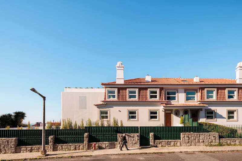Street view of white corrugated metal volume adjoining red tile roof townhouse with stone wall base