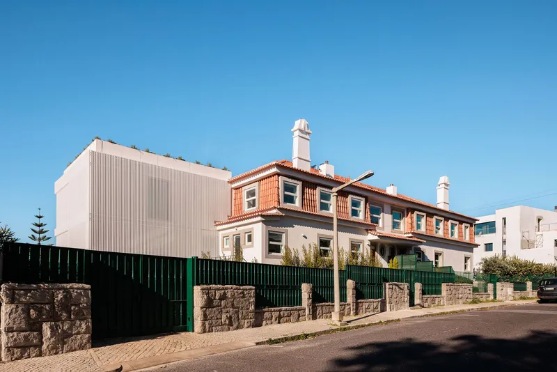 White ribbed facade addition beside traditional terrace housing with green metal fence along cobbled street