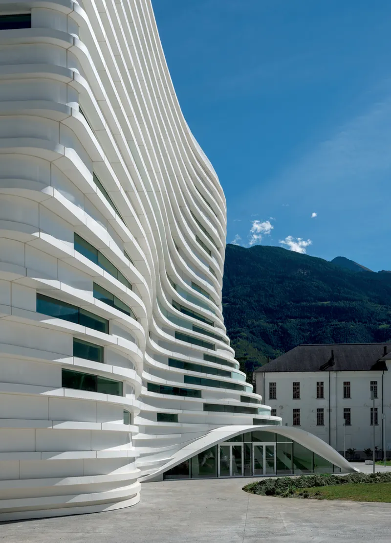 Layered white horizontal facade wrapping around a glass entrance pavilion with mountains beyond