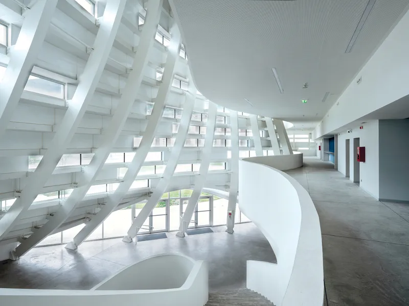 Interior corridor with angled white structural ribs along glazed wall and curved balustrade in concrete