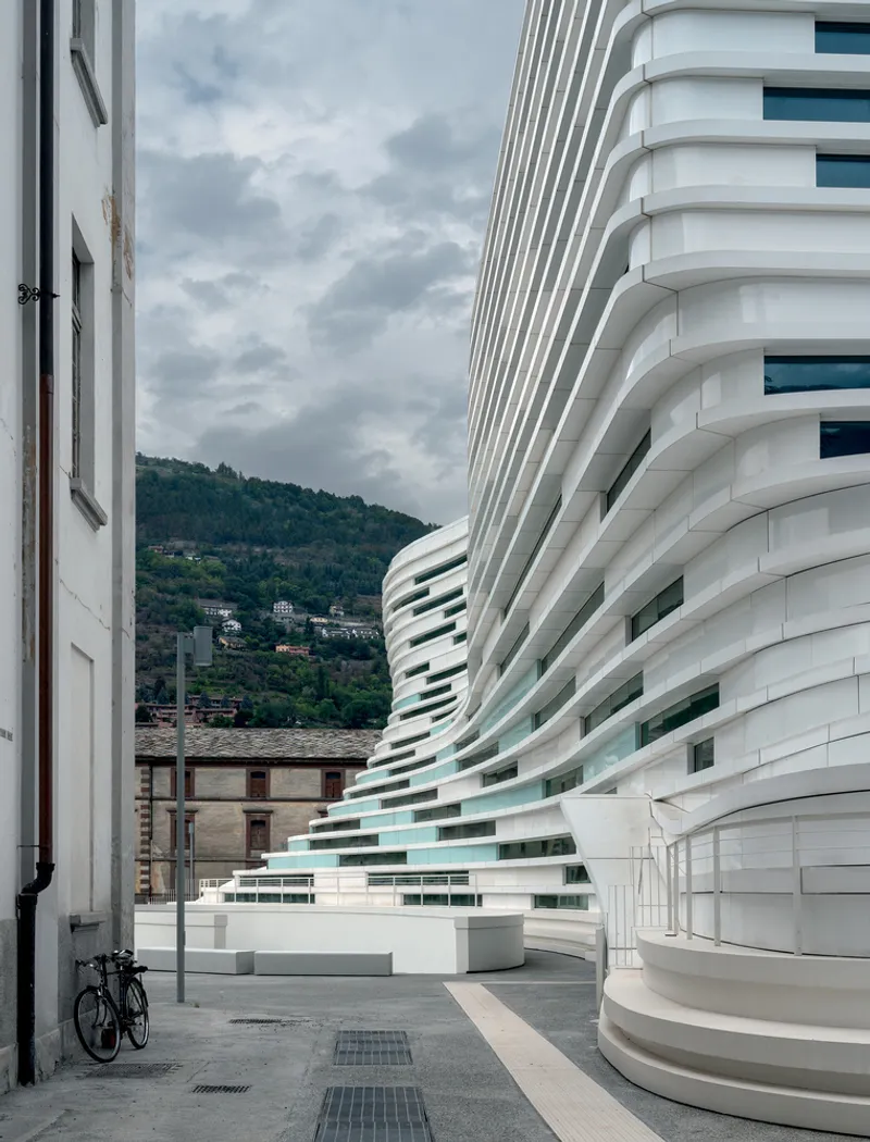 Curved facade with horizontal white bands meeting an older building under an overcast sky