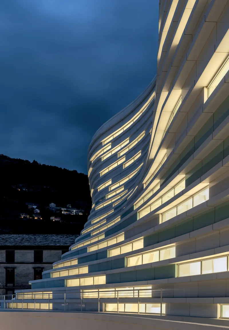 Illuminated facade with horizontal bands of glazing at dusk against a hillside backdrop