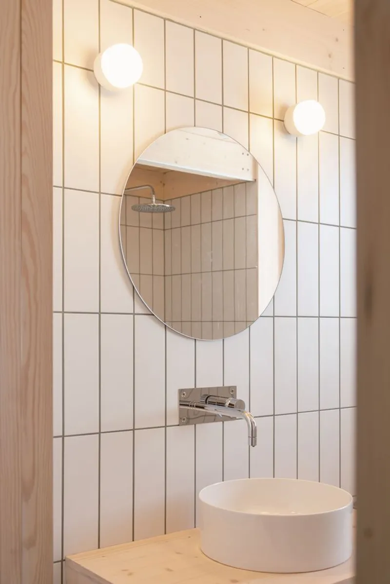 Bathroom vanity with round mirror against white vertical tile and globe sconces flanking the sink