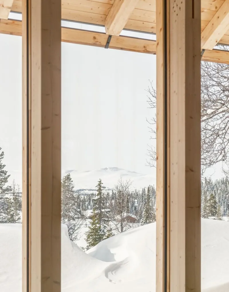 Floor-to-ceiling window framed by timber columns and rafters overlooking snow-covered forest and distant mountains