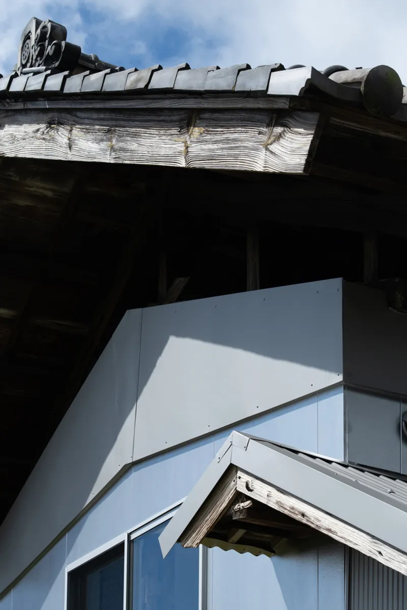 Upward view where the pale blue cladding meets weathered timber eaves and curved clay roof tiles