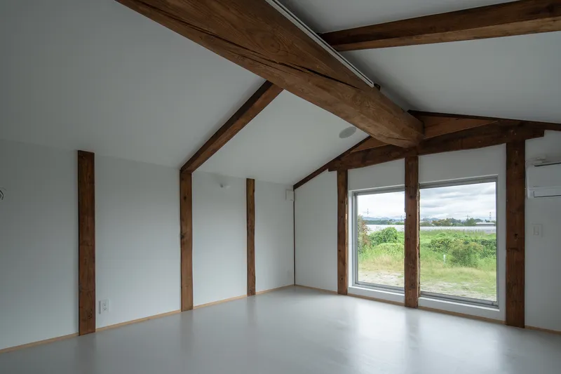 Interior room with exposed timber beams and columns framing a large window overlooking a grassy meadow