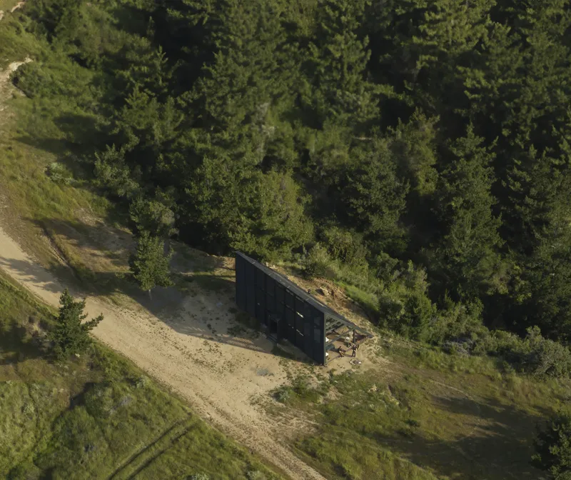 Aerial view of a dark rectangular cabin on sandy ground surrounded by dense forest