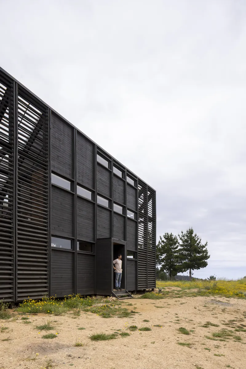 Black louvered facade with gridded windows facing sandy terrain and a distant tree under overcast skies