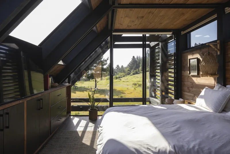 Bedroom with sloped timber ceiling, black steel frame glazing, and morning sunlight across white bedding