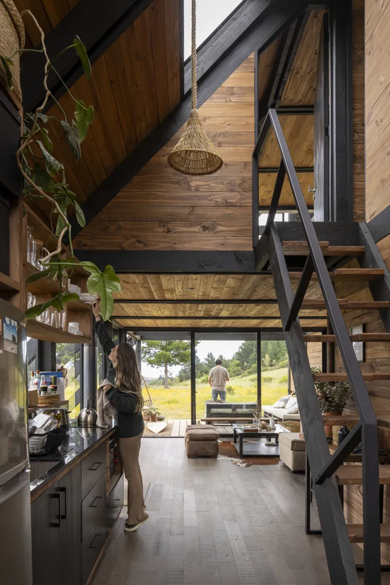 Double-height interior with timber walls, a steel ladder, kitchen below and mezzanine above overlooking landscape