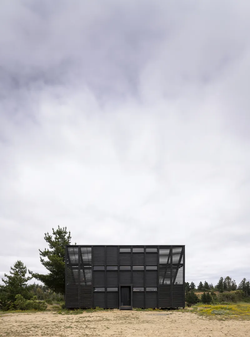 Black corrugated metal facade with horizontal louvers and windows on sandy ground under overcast sky