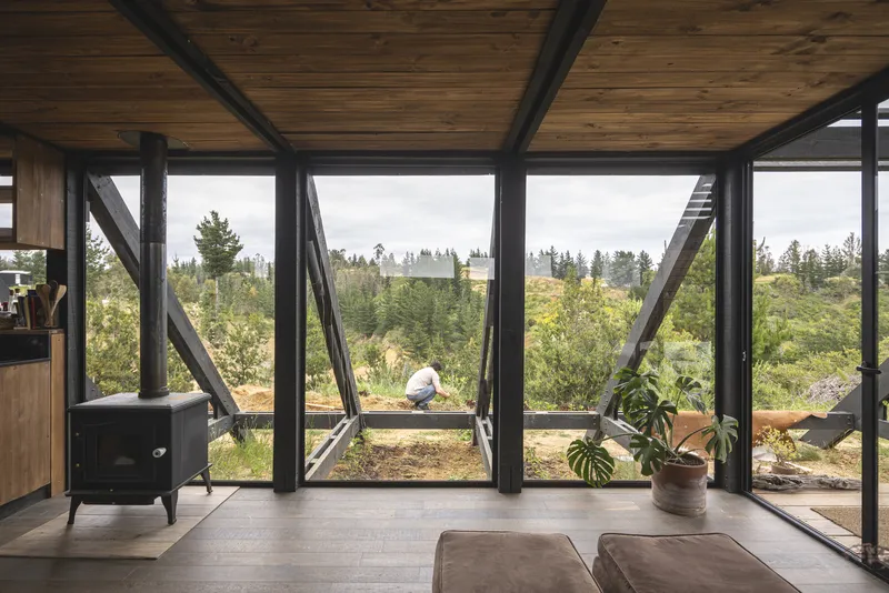 Interior living space with full-height angled glazing, exposed timber ceiling and person seated outside