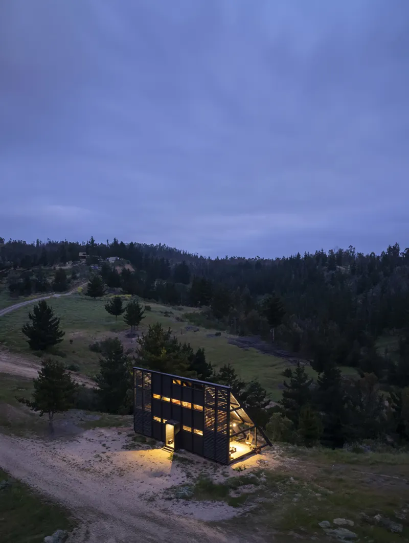 Aerial view of illuminated A-frame cabin on hillside meadow surrounded by forest at twilight