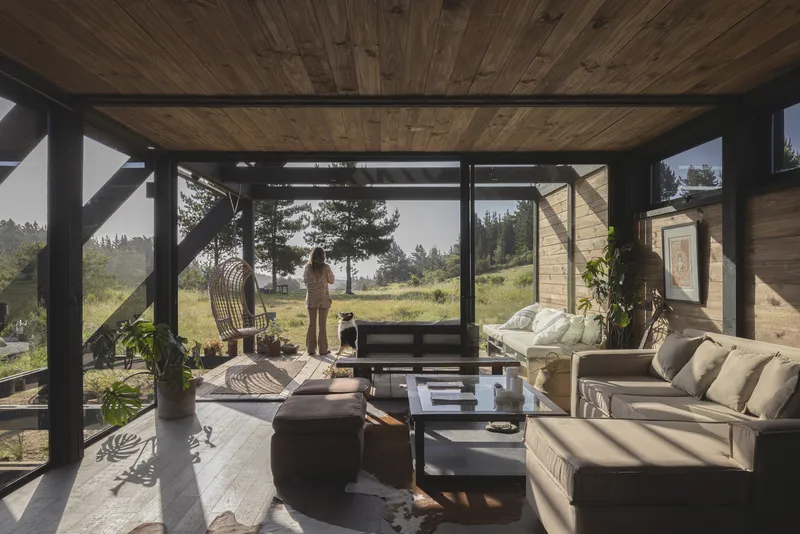 Living area with timber ceiling and black steel frame opening to grassland with figure standing outside