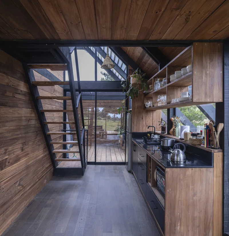 Galley kitchen with timber shelving and black steel ladder ascending beneath diagonal exposed bracing