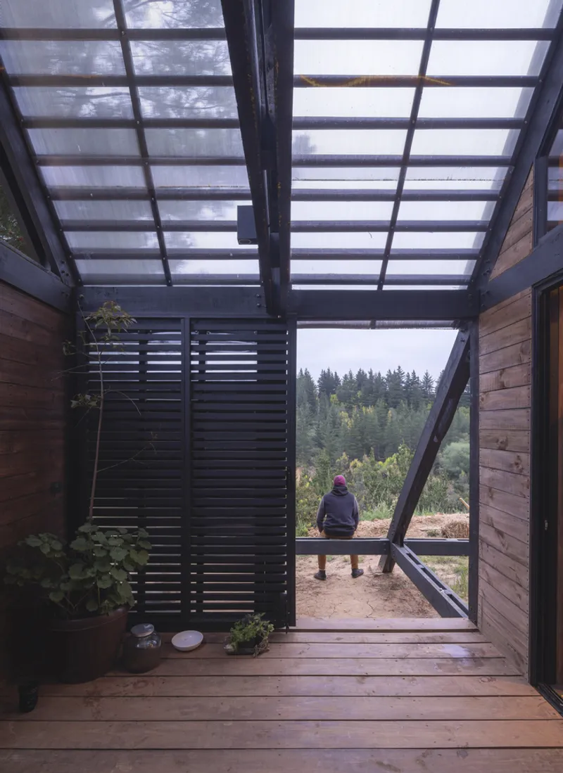Covered timber deck with translucent roof panels and seated figure overlooking forested hillside