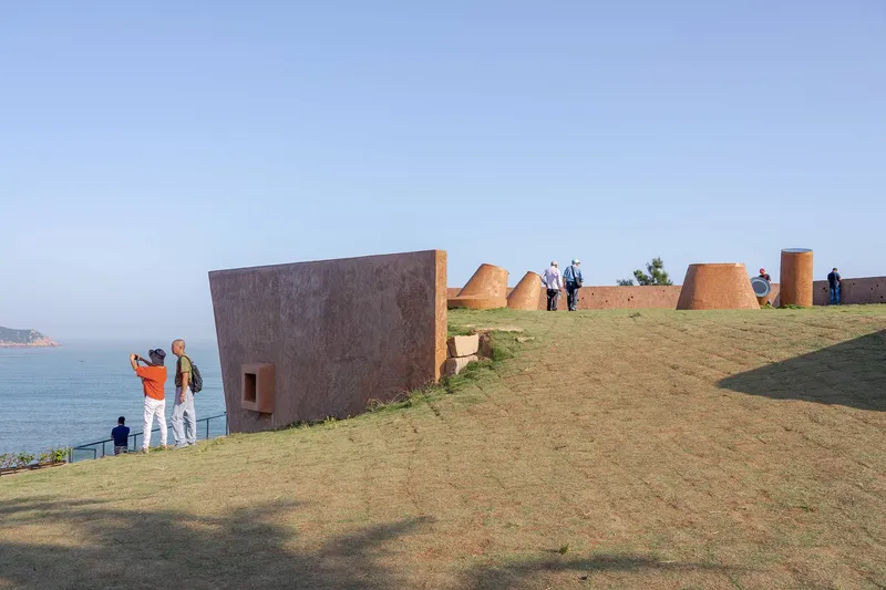 Tilted rammed-earth volume and cylindrical forms scattered across a grassy clifftop overlooking the sea