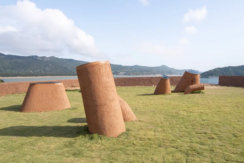 Angled cylindrical rammed-earth forms rising from a mown lawn with mountains and water beyond