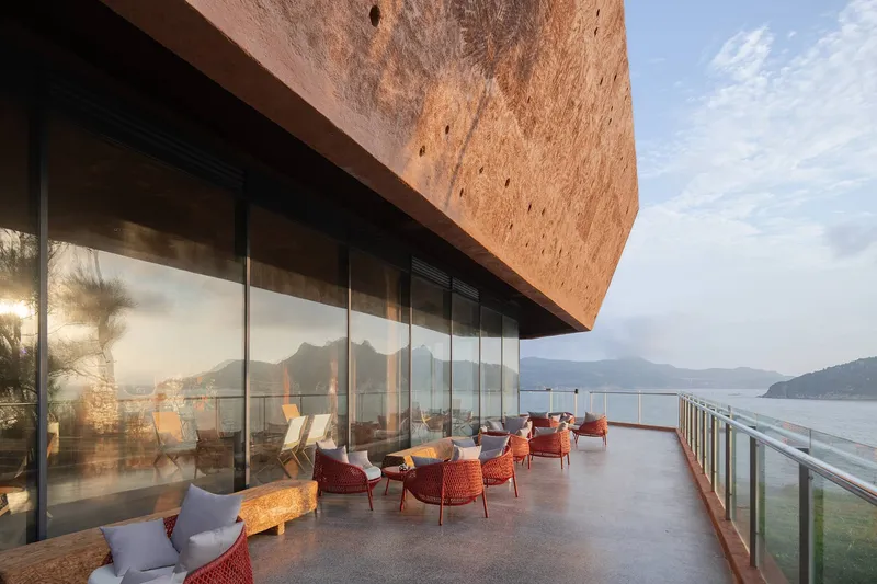 Glass-enclosed terrace beneath a tilted rammed-earth volume with seating overlooking the coastline at dusk