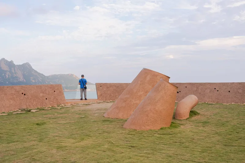 Terracotta-finished sculptural forms scattered across a grass lawn with a visitor and lake in the background