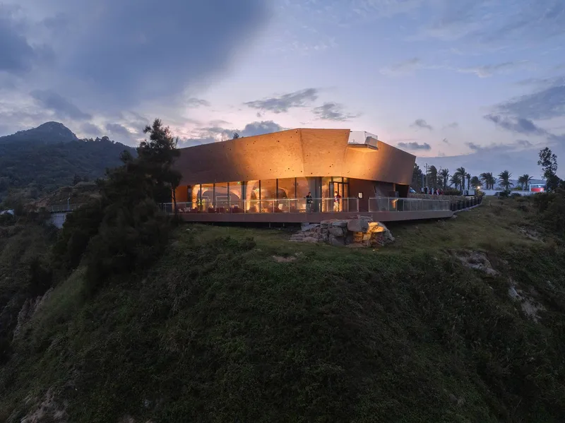 Curved rammed earth pavilion perched on a hillside at dusk with interior lights glowing