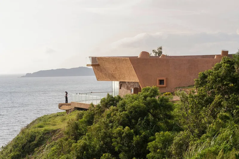 Cantilevered rammed earth roof extending over a coastal terrace with a person standing at the glass railing