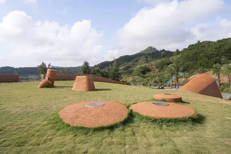 Circular rammed earth platforms and tilted forms arranged on a lawn with mountains in the distance