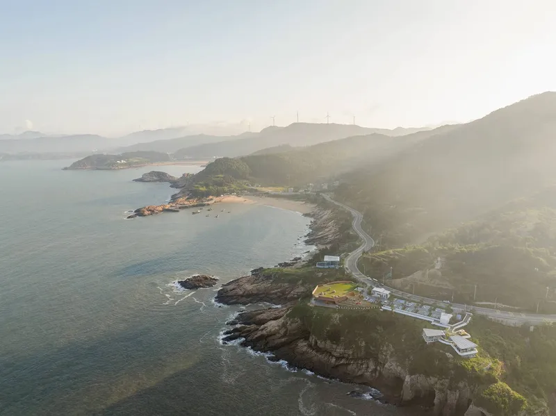 Aerial view of coastal promontory with low buildings nestled along a winding road in morning haze