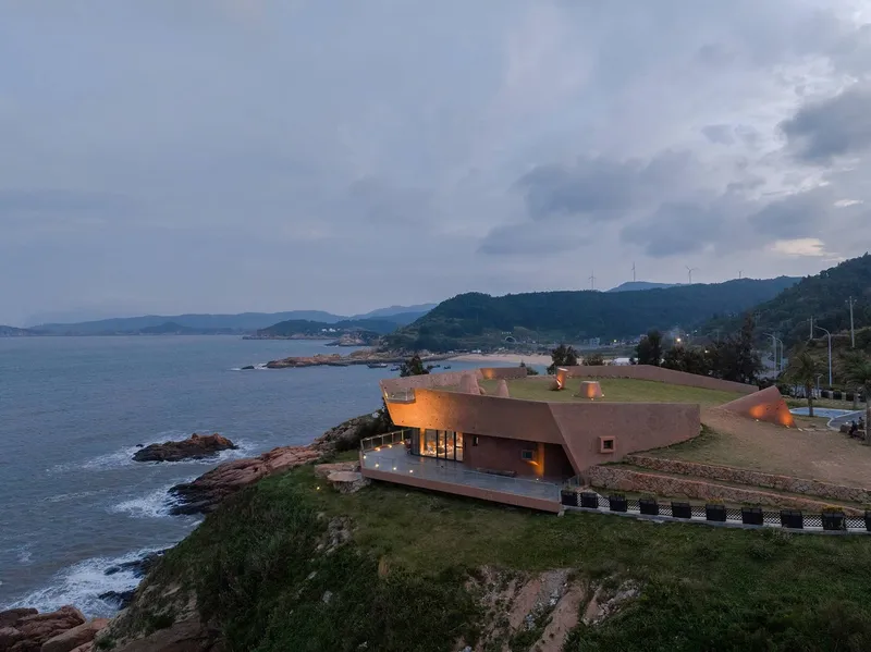 Low-slung structure with angular roof planes perched on rocky coastline under stormy evening sky