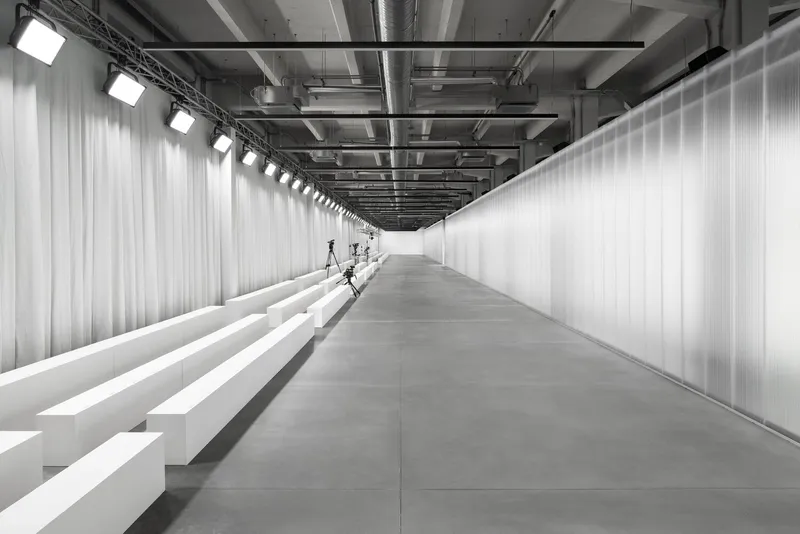 Long interior corridor with white tiered seating along translucent curtain walls and exposed truss ceiling