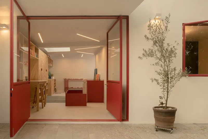 View through red metal frame doorway into studio space with plywood storage and a potted olive tree