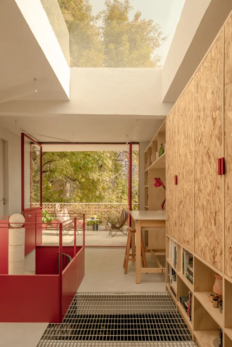 Interior corridor with red metal partitions and black grating floor beneath a skylight framing foliage