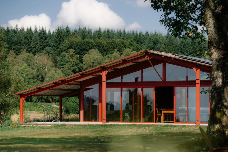 Red timber-framed pavilion with glass-walled facade and overhanging corrugated roof set against a forested hillside