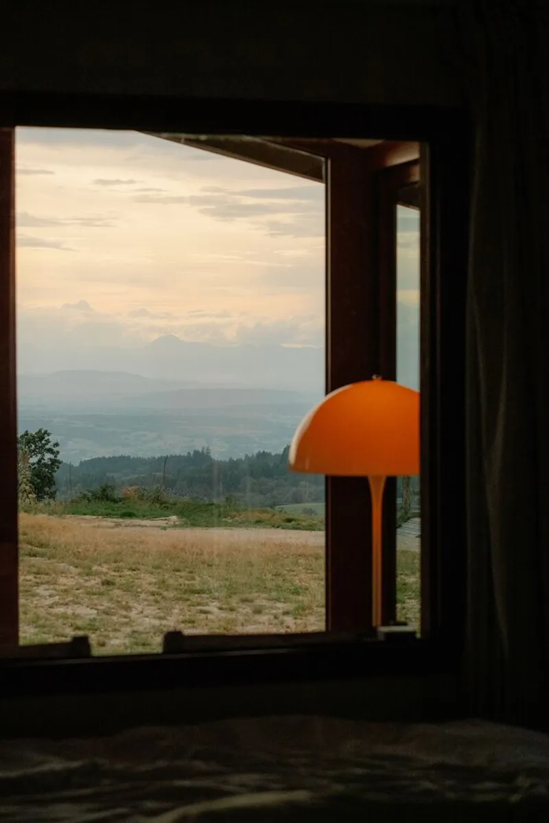 Interior view through timber-framed opening with orange pendant lamp toward forested hills at dusk