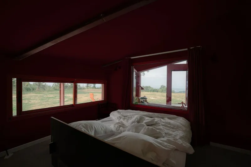 Bedroom with red walls and corner windows framing dry grassland under overcast sky